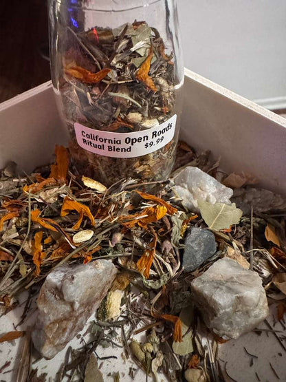 A glass jar labeled 'California Open Roads Ritual Blend $9.99' filled with dried herbs and crystals, surrounded by loose botanicals and stones inside a white box.