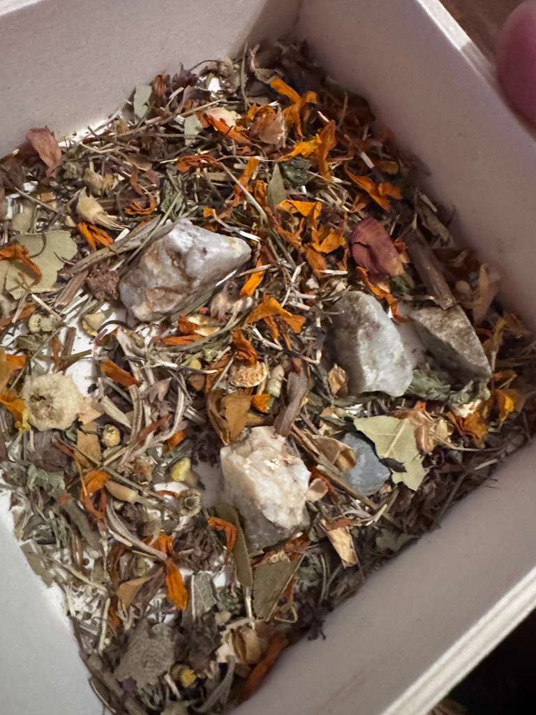 A close-up view of a white square container filled with a loose herbal and mineral blend, featuring dried orange flowers, green leaves, brown stems, and several pale gray to off-white stones.