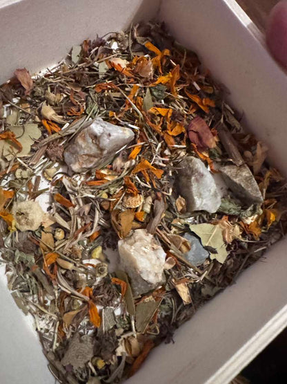 A close-up view of a white square container filled with a loose herbal and mineral blend, featuring dried orange flowers, green leaves, brown stems, and several pale gray to off-white stones.