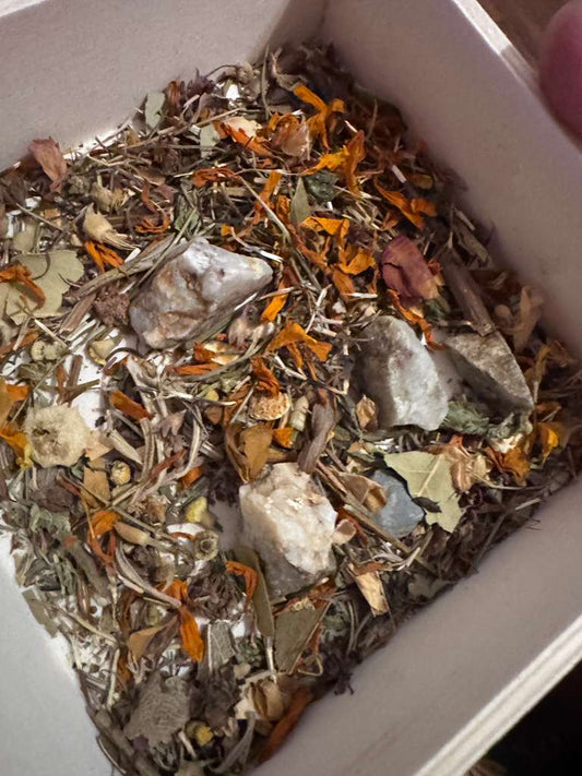 A close-up view of a white square container filled with a loose herbal and mineral blend, featuring dried orange flowers, green leaves, brown stems, and several pale gray to off-white stones.
