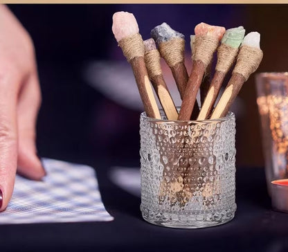 A hand holds a small textured glass cup filled with wooden crystal pens wrapped in twine and topped with colorful chakra stones, set against a dark background with a blurred candle and text overlay at the bottom.
