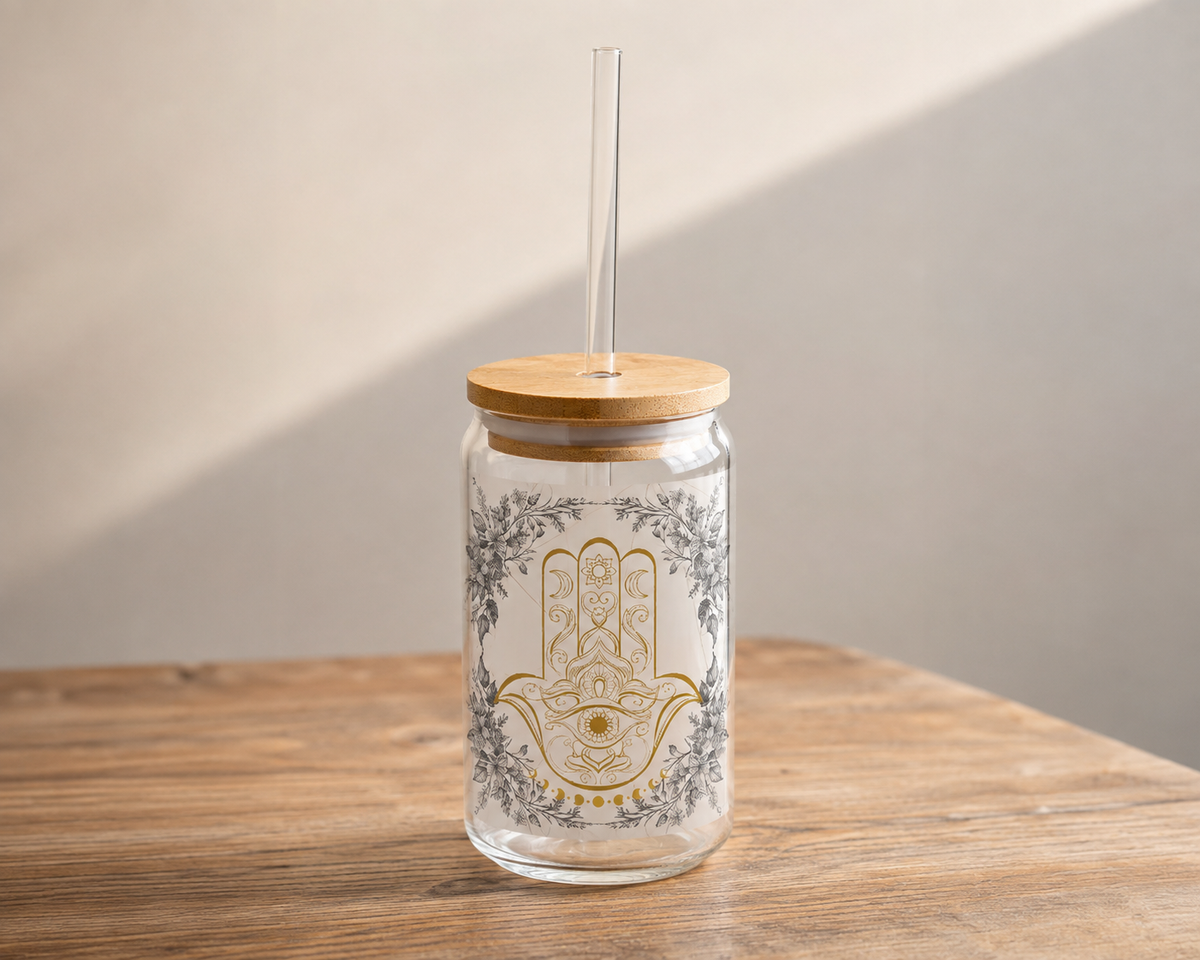 A clear glass tumbler with a bamboo lid and clear straw, featuring a gold and gray Hamsa hand design on the front, set on a wooden table against a neutral wall.