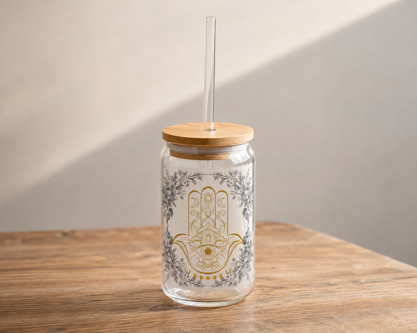 A clear glass tumbler with a bamboo lid and clear straw, featuring a gold and gray Hamsa hand design on the front, set on a wooden table against a neutral wall.