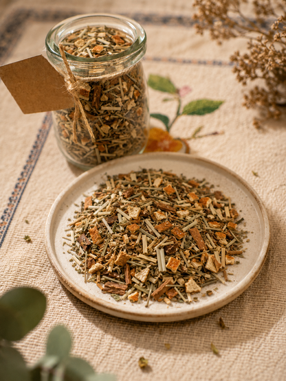 A glass jar filled with loose herbal tea or botanical blend sits behind a ceramic plate piled with the same dried tea mixture. The scene is styled on a textured beige fabric with dried flowers and green leaves around it.
