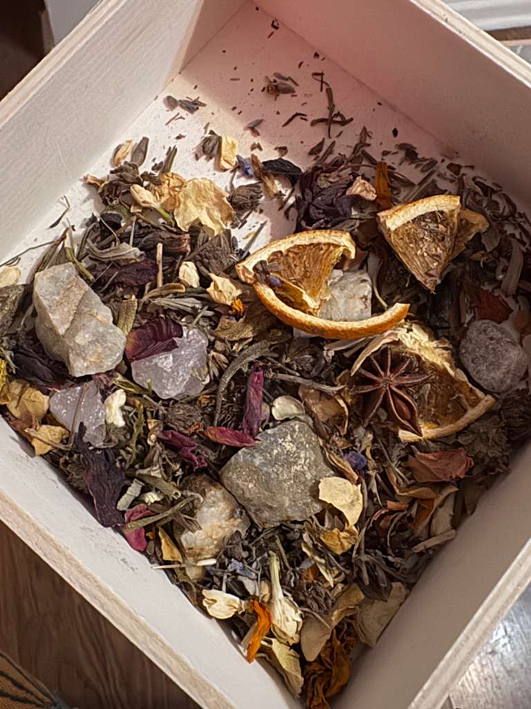 A close-up view of a dried botanical blend inside a light-colored wooden box. The blend includes dried orange slices, star anise, purple and white flowers, and various natural stones.
