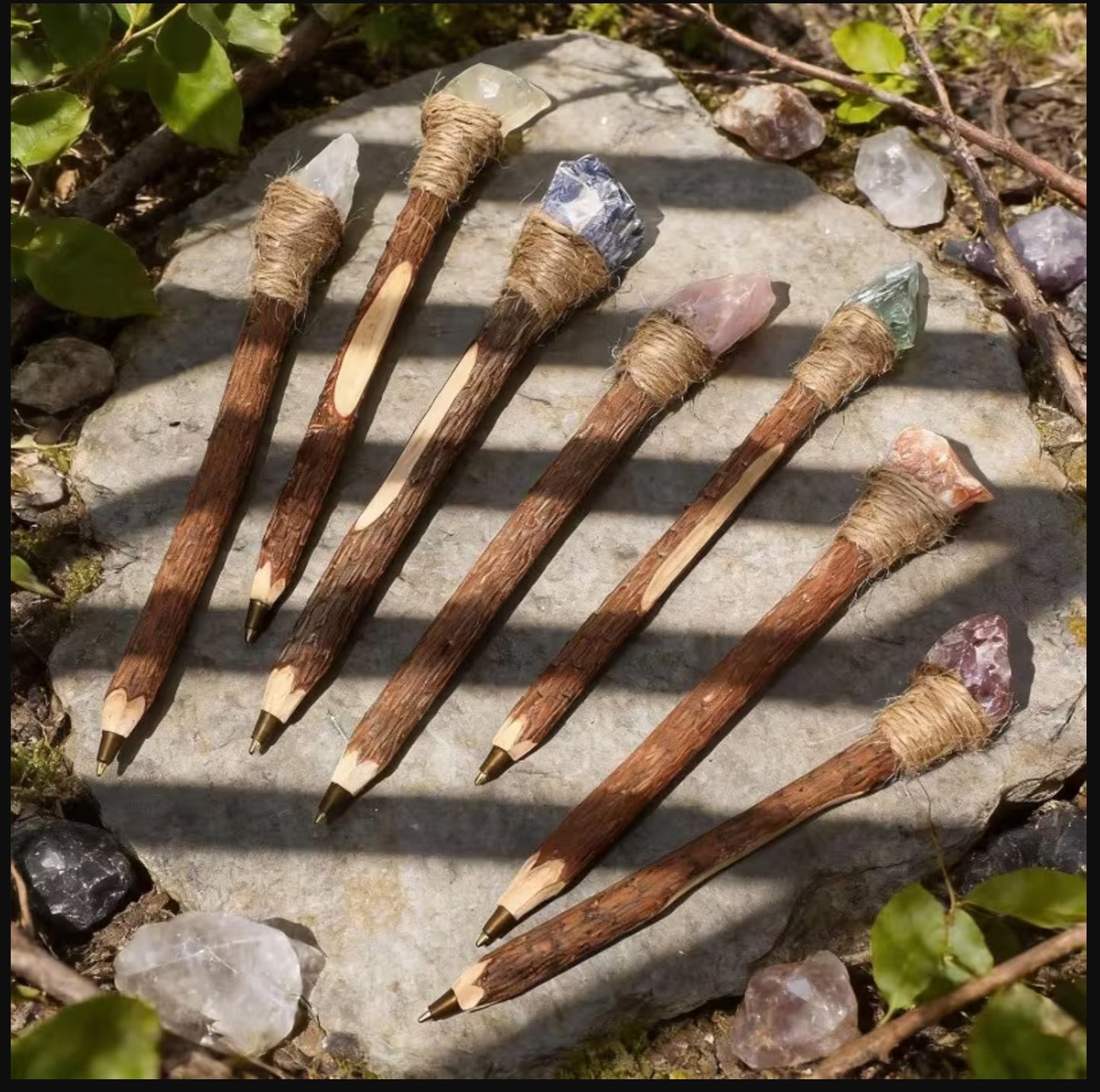 Seven handmade wooden pencils with natural animal-hide wrapped ferrules and colorful crystal tips, arranged on a flat stone outdoors with sunlight and shadows.