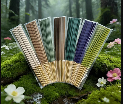 Seven packaged incense sticks in different colors (beige, brown, green, teal, blue, yellow-green) arranged in a fan shape on mossy forest ground with small flowers.